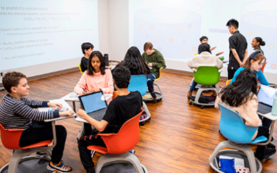 Students sit in rolling desks and chairs in a science class with computers with walls that are filled with screens displaying information about their chemistry studies.